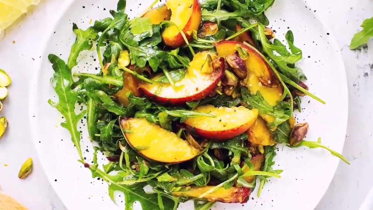 Đĩa salad rau rocket, quả đào và hạt dẻ cười trên nền đĩa trắng.