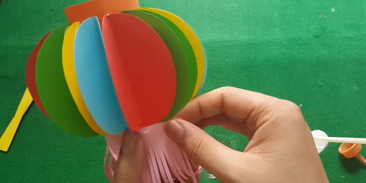 Dán hai cạnh chiều rộng hình chữ nhật đã dán những hình tròn lại, lấy miếng tua rua vừa cắt dán vào đáy của lồng đèn